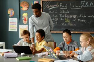 A teacher standing behind children sitting at a table looking at laptops.